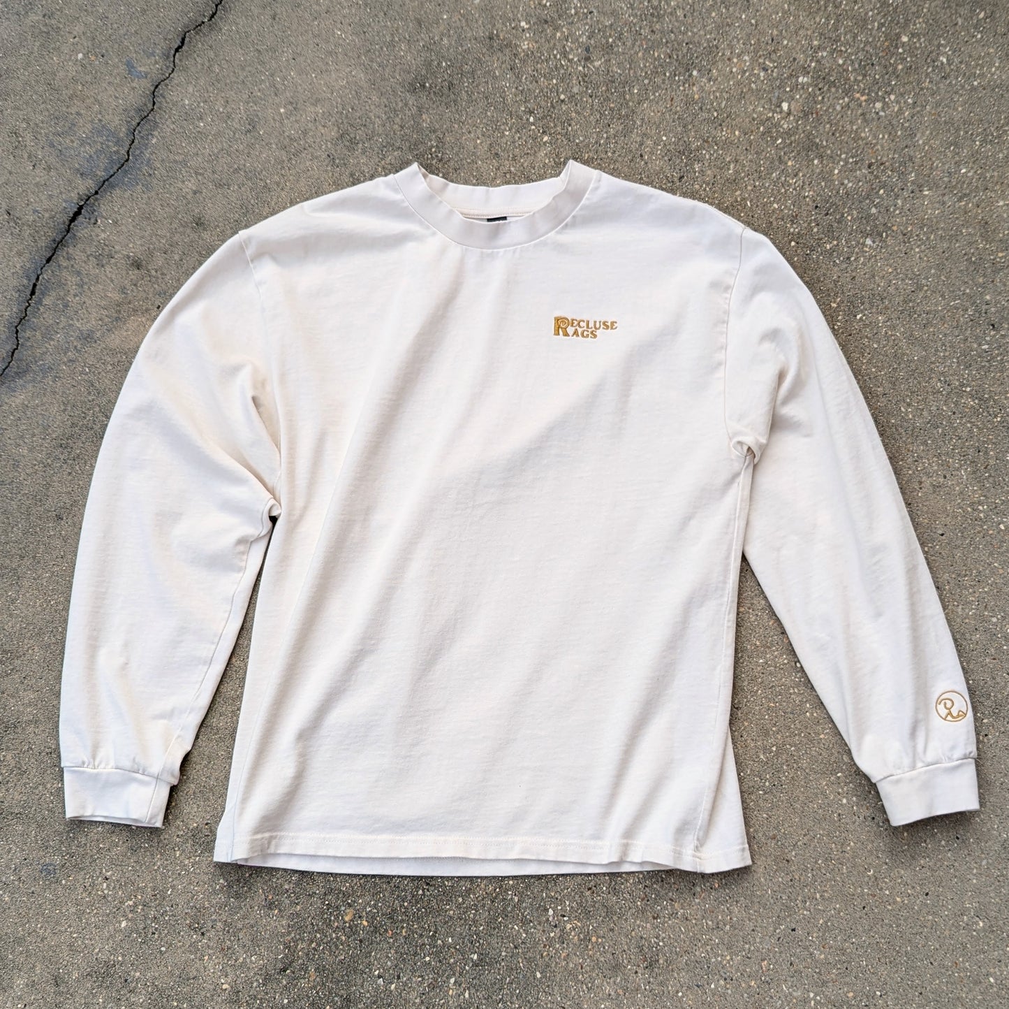 Oversized, long sleeve t-shirt in ecru color laid out on concrete, with Recluse Rags embroidered on left chest and logo on left sleeve. All embroidery in gold.