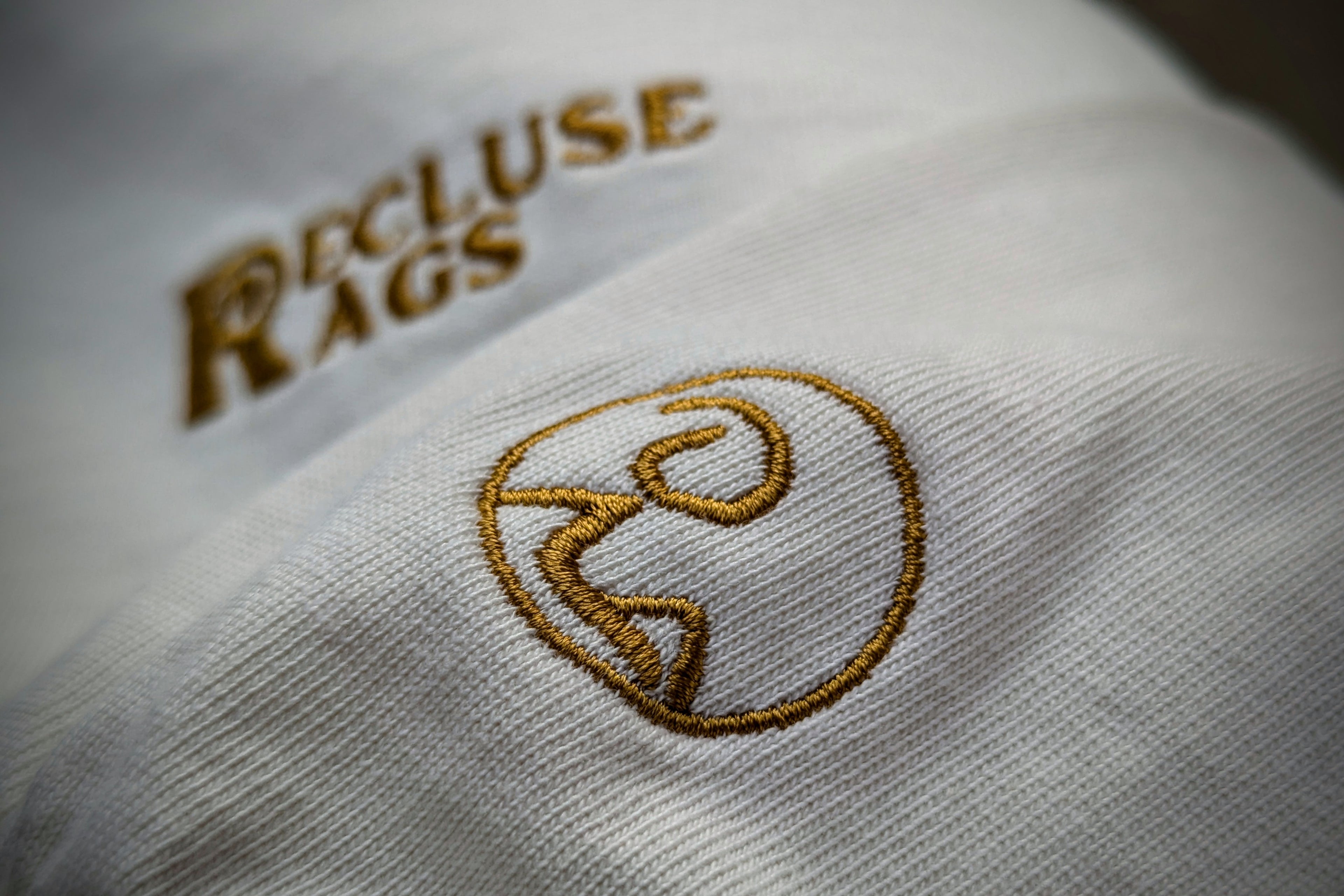 Dramatic zoomed in view of embroidered logo on left sleeve of Ecru colored Ultra-Heavy Recluse Rags Long Sleeve Tee.
