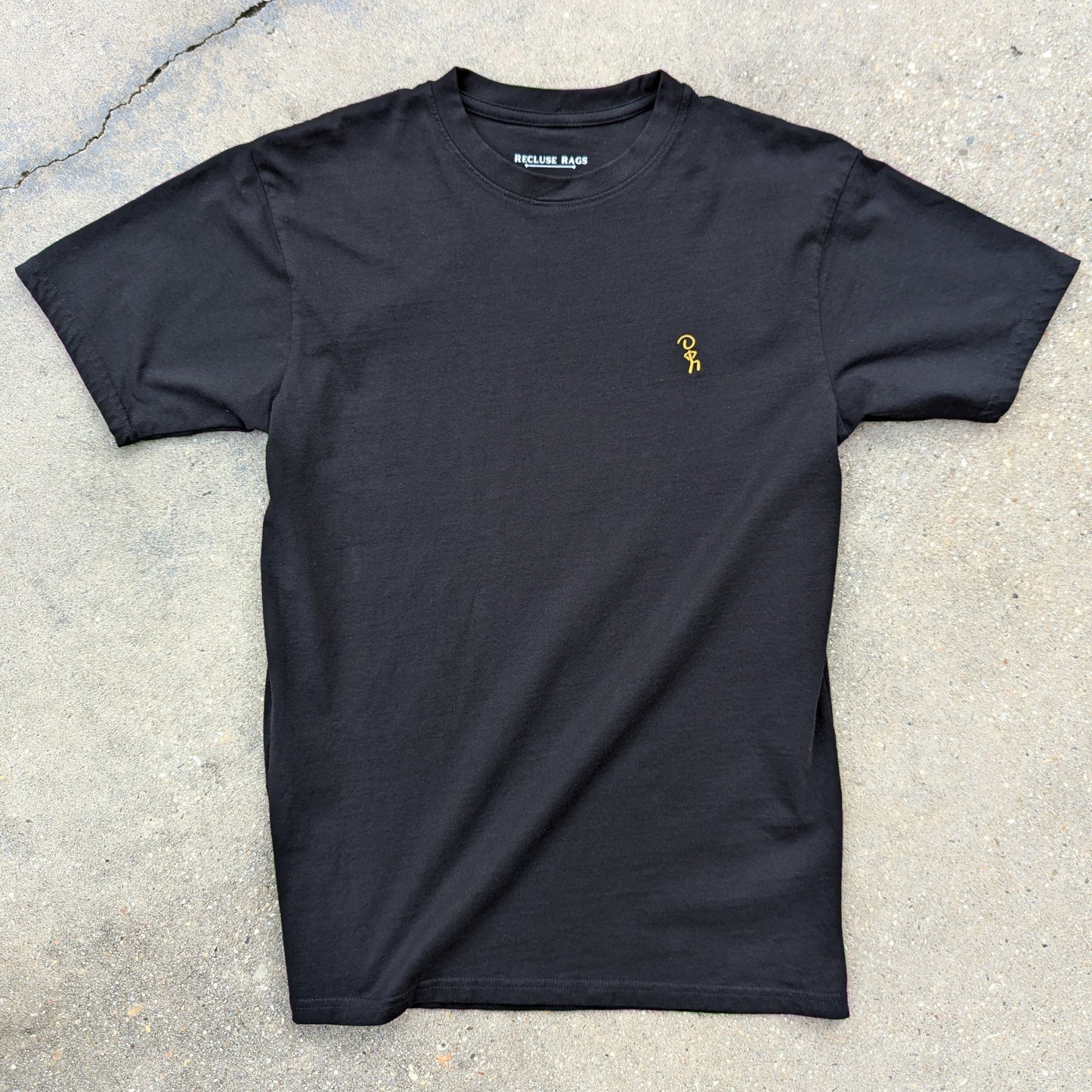 Gold embroidered Jaunty Man on premium black t-shirt. Laid out on concrete.