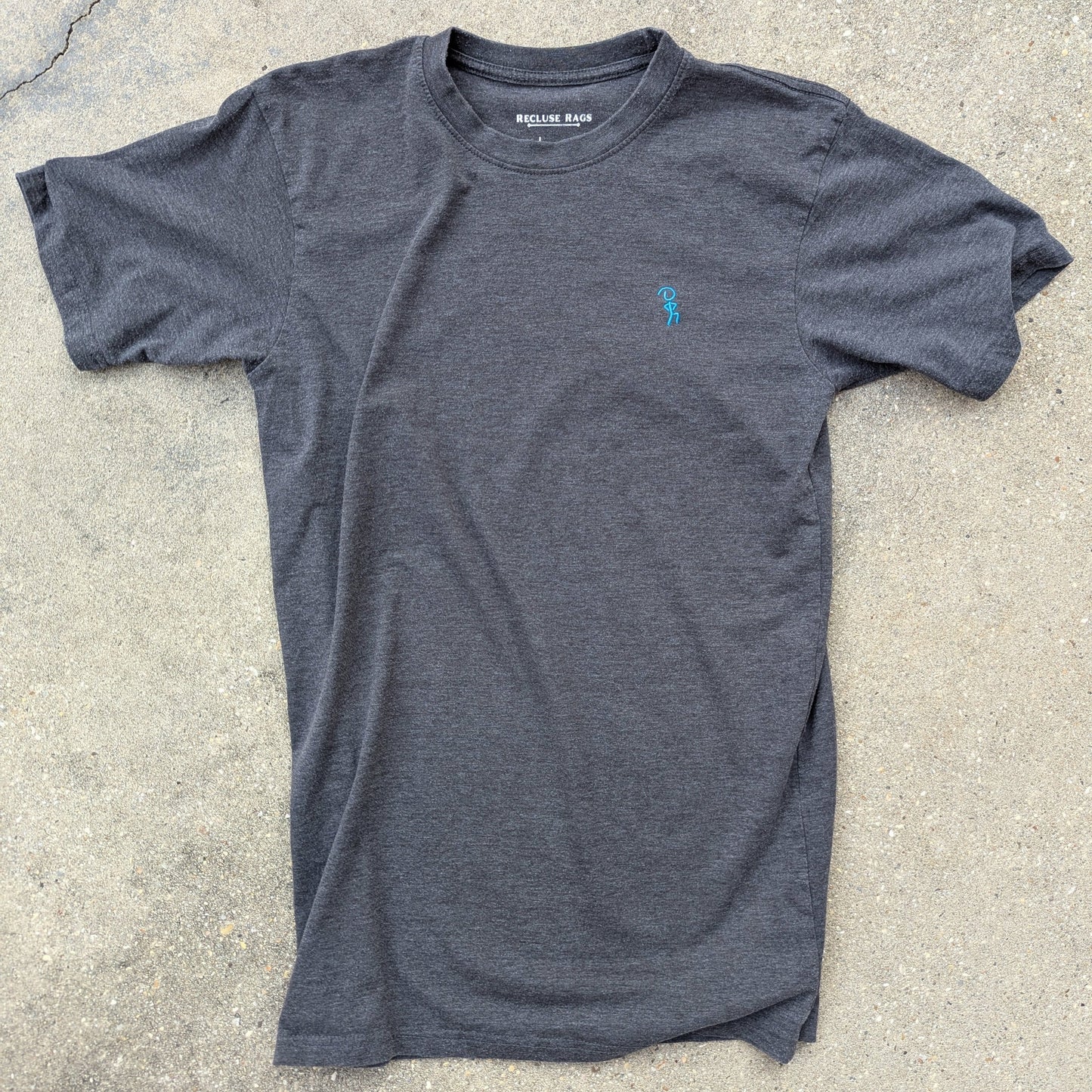 Blue embroidered Jaunty Man on premium charcoal t-shirt. Laid out on concrete.