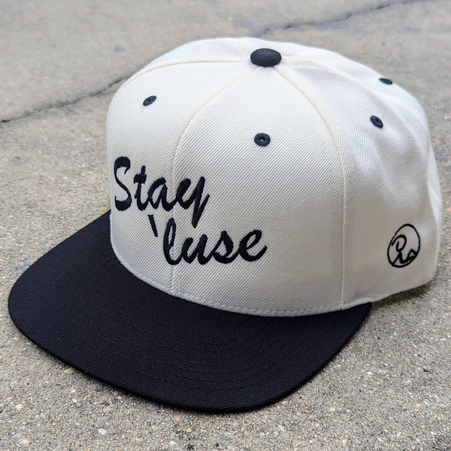 Stay 'luse Snapback in black and natural. Stay 'luse embroidered across the front in black. Recluse Rags logo without text visible embroidered on side of hat.