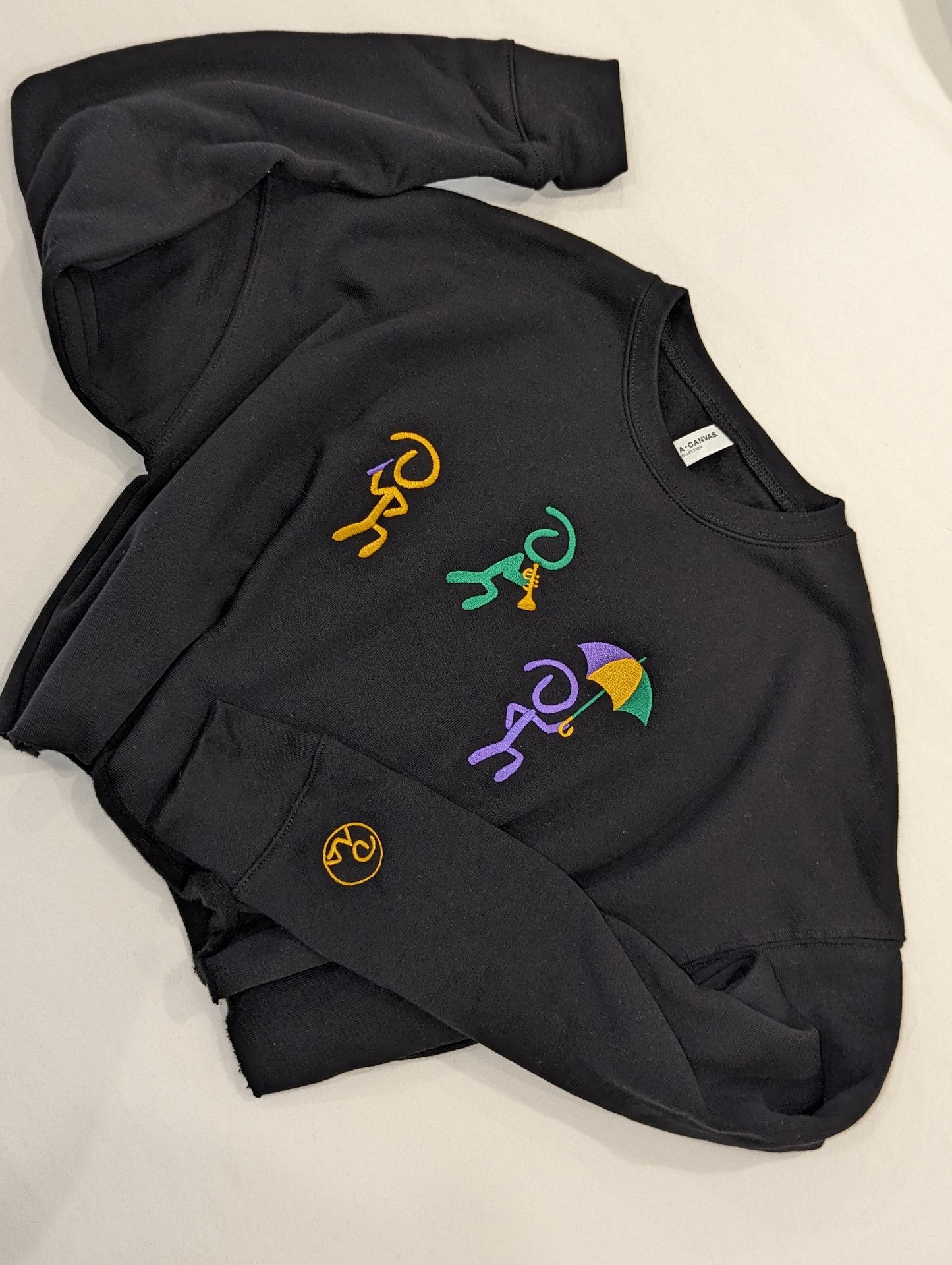 Black cropped sweater with 3 "Jaunty Man" stick figures embroidered in gold, green, and purple, and holding a drink, a trumpet, and an umbrella, respectively.