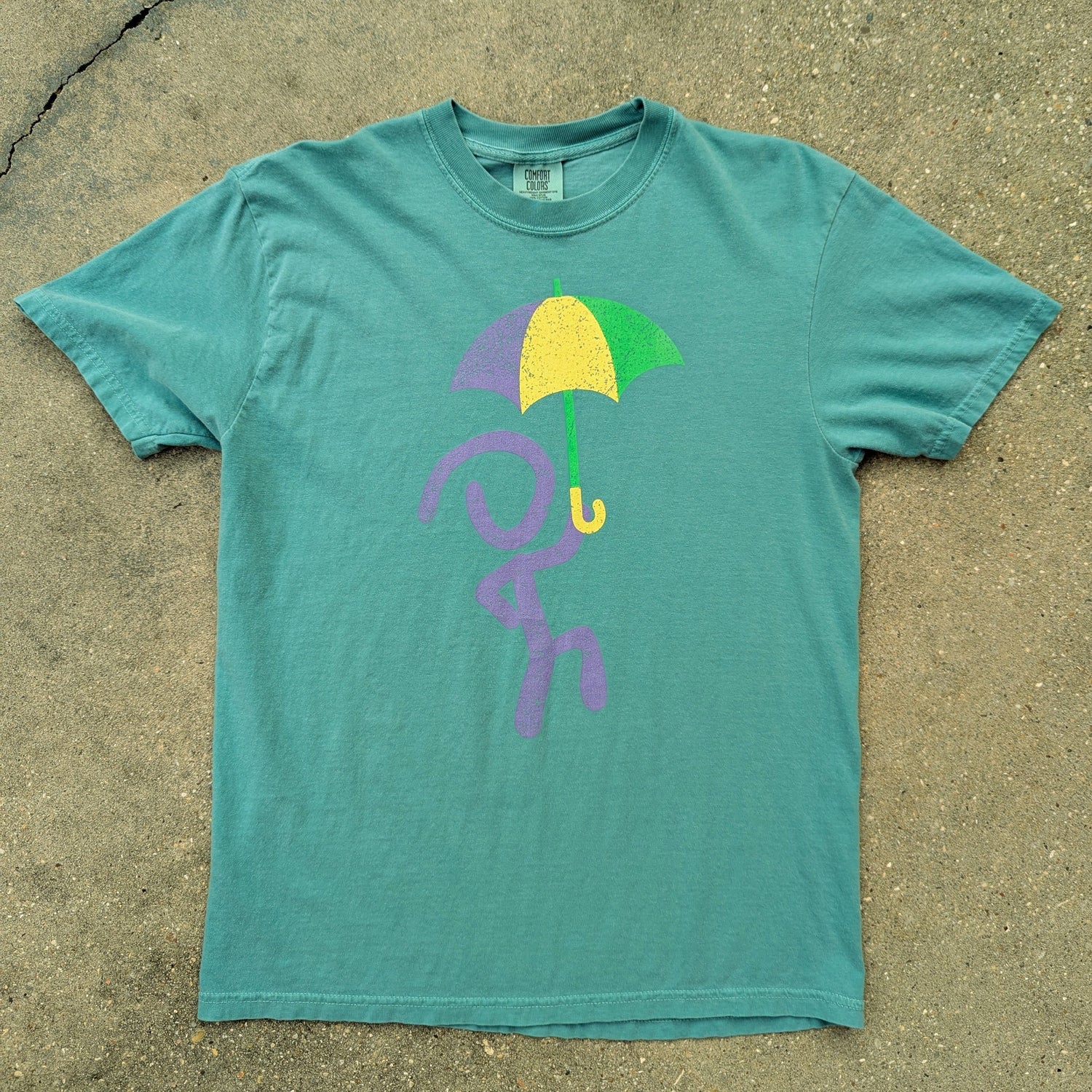 Jaunty Man in purple holding up a purple, green, and gold umbrella on seafoam t-shirt. Laid out on concrete.