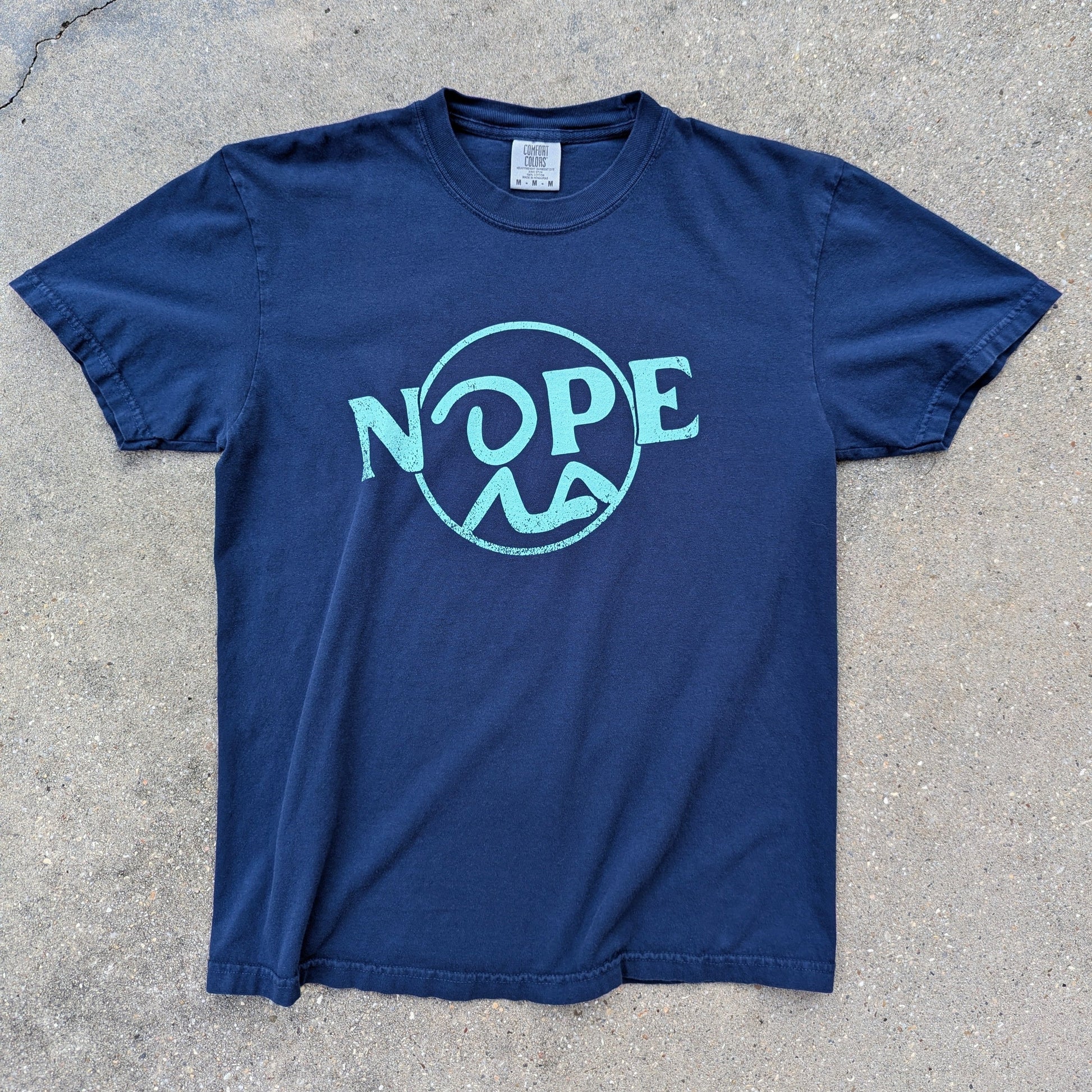 Recluse Rags Nope logo in cyan on Navy t-shirt. Laid out on concrete.