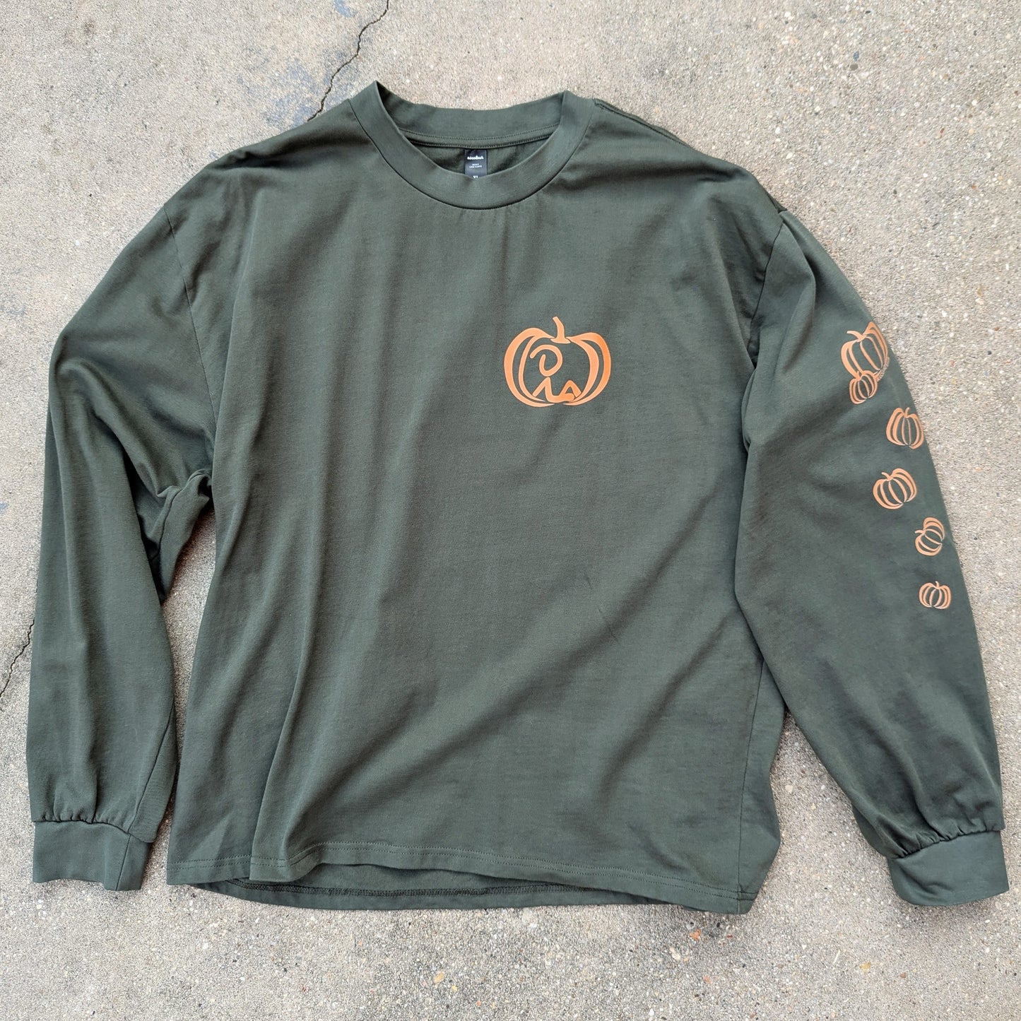 Oversized, long sleeve t-shirt in cypress color laid out on concrete, with Jaunty Man reclined inside pumpkin on left chest and varied pumpkins down left sleeve. all colored a rusty orange shade.