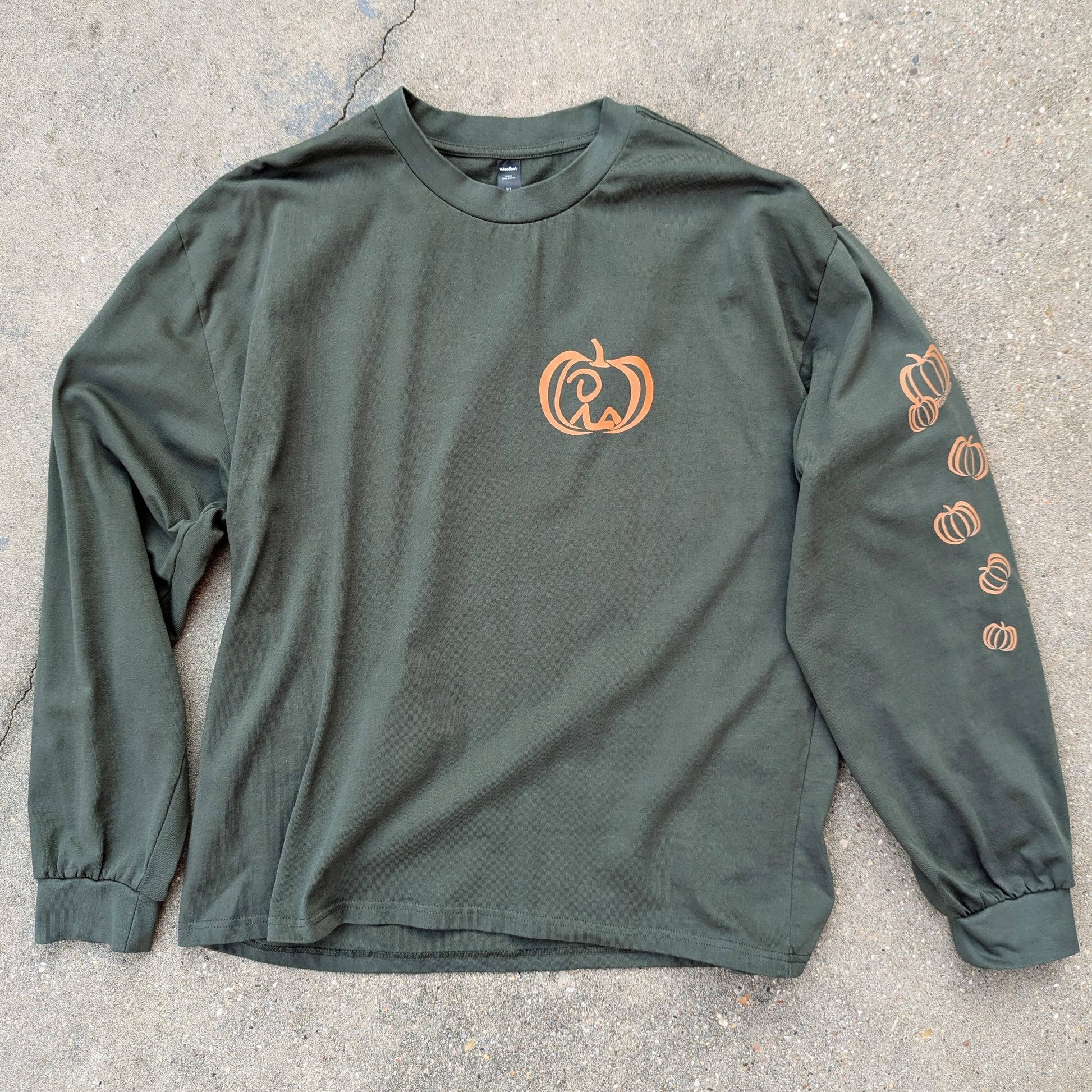Oversized, long sleeve t-shirt in cypress color laid out on concrete, with Jaunty Man reclined inside pumpkin on left chest and varied pumpkins down left sleeve. all colored a rusty orange shade.