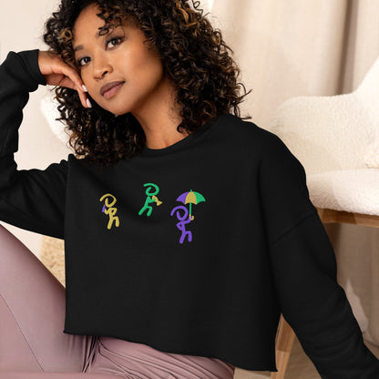 Woman posing in black cropped sweater with 3 "Jaunty Man" stick figures embroidered in gold, green, and purple, and holding a drink, a trumpet, and an umbrella, respectively. Recluse Rags logo without text on left wrist.